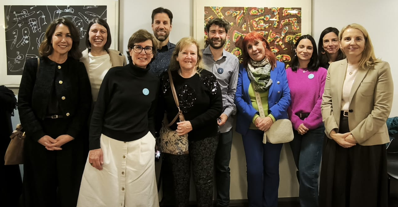 Un grupo de diez adultos posan y sonríen en el interior delante de una obra de arte enmarcada; la mayoría están de pie, muy juntos, con ropa informal o semiformal.