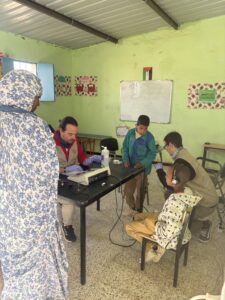 Dos cooperantes se sientan a la mesa de un aula y atienden a tres niños y a una mujer que se encuentra cerca. El equipo médico está sobre la mesa, y la habitación tiene paredes verdes con decoraciones de colores.