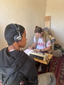 Una mujer escribe en un portapapeles en una pequeña mesa con equipo de audio mientras un chico con auriculares está sentado cerca. Parecen estar en una habitación modesta y escasamente amueblada.