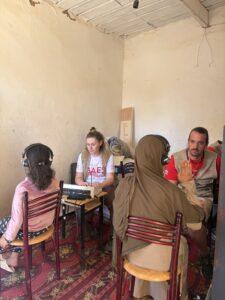Dos cooperantes con uniformes blancos y rojos se sientan ante una mesa con equipo de audio y hablan con dos mujeres locales con pañuelos en la cabeza y auriculares, en el interior de una sencilla habitación con paredes color crema y una alfombra estampada.